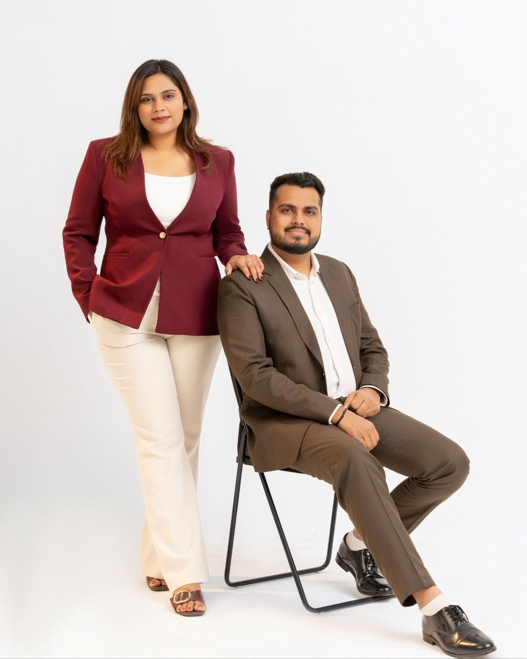 Riddhi standing in a maroon blazer beside Mann seated in a brown suit, photographed in the studio